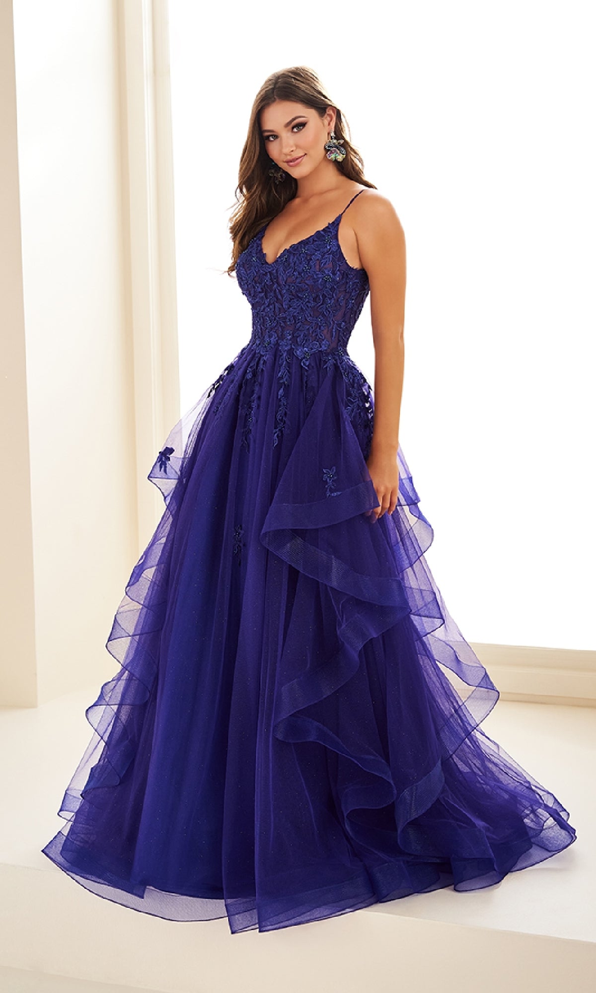 A woman wears the Ellie Wilde EW36085 long prom dress indoors, featuring deep blue glitter tulle, a fitted lace-embroidered corset bodice, and a layered tulle skirt.